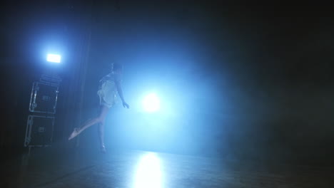 a-young-girl-in-a-white-dress-dances-a-modern-ballet-spins-on-the-leg-and-jumps-on-the-stage-with-smoke-against-the-blue-spotlights.